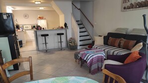 Ein Wohnzimmer mit einer Couch, einem Couchtisch und einem Sessel. Es gibt eine Küchenzeile mit einem Kühlschrank, einer Mikrowelle und einem Barhocker. Eine Treppe führt zu einer oberen Ebene.