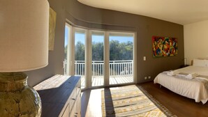 Un dormitorio con una cama grande, una ventana con vistas a árboles, un cuadro colorido en la pared y una planta en maceta.