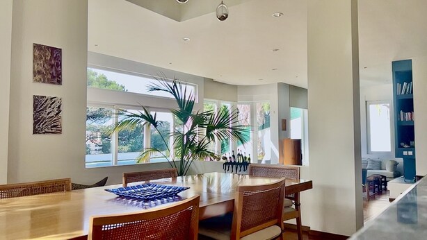 Un comedor moderno con una mesa y sillas de madera, ventanas amplias con vistas a zonas verdes y una estantería con libros.