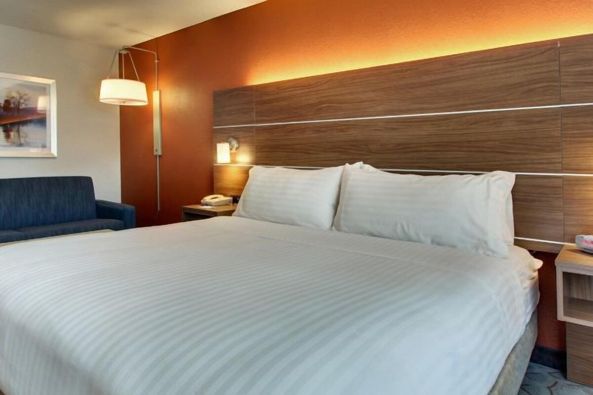 A hotel room with a large bed, a desk, a chair, and a nightstand with a lamp.