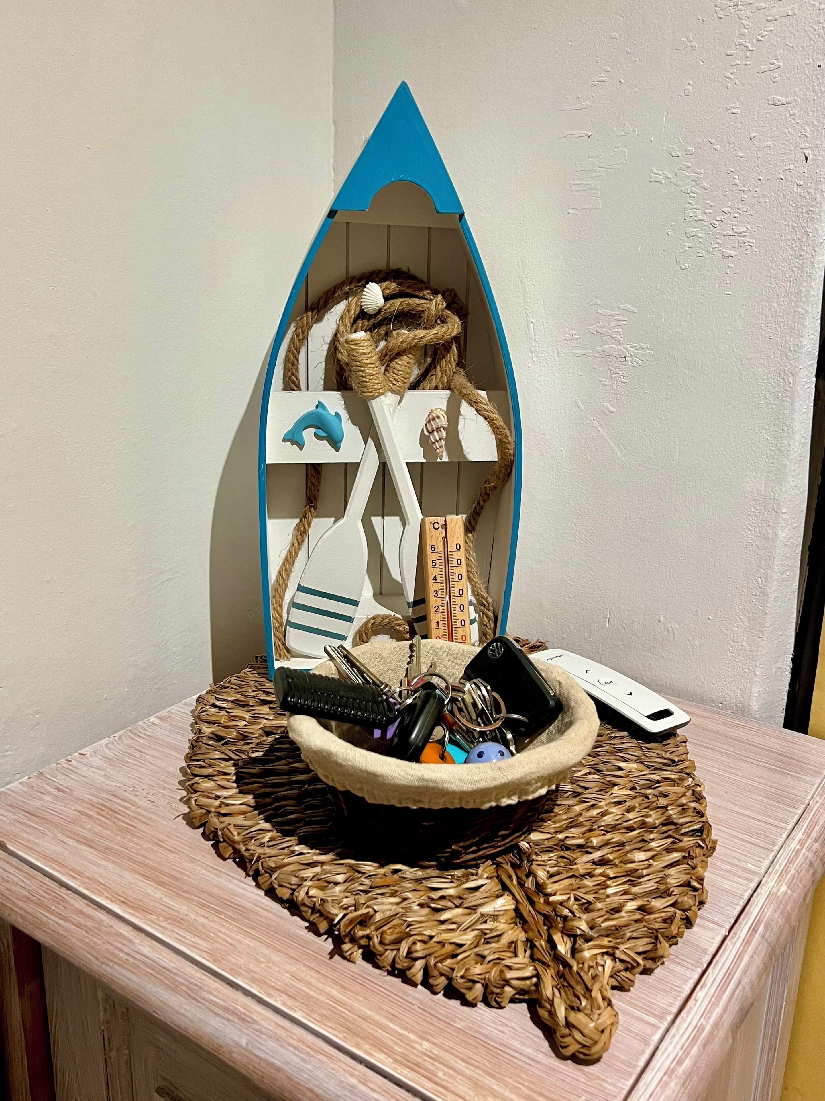 Una mesita pequeña con un protector tejido, un estante decorativo con forma de barco y una cesta con llaves y un mando a distancia.
