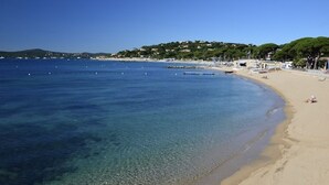 Ein Strand mit klarem, blauem Wasser, sandigem Ufer und Gebäuden im Hintergrund.