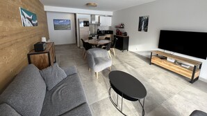 En moderne stue med en grå sofa, et lille sort bord, et fjernsyn og et køkken i baggrunden.