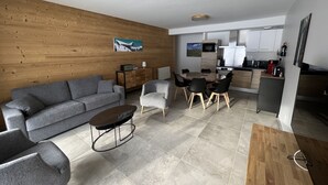 En moderne stue med en grå sofa, et sofabord og et spiseområde med et træbord og stole. Et køkken med skabe og hårde hvidevarer er synligt i baggrunden.