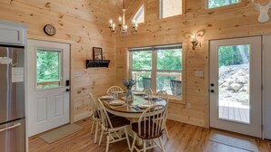 Interior de una cabaña de madera con una mesa de comedor para cuatro personas, una araña y un ventanal con vistas a un bosque.