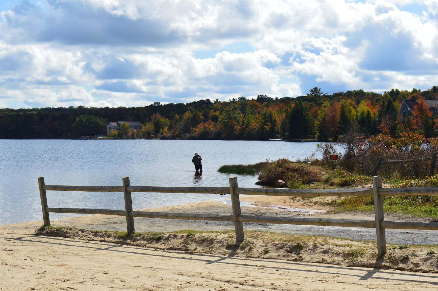 A wooden fence overlooking a lake with a person fishing, surrounded by autumn-colored trees and a clear sky.