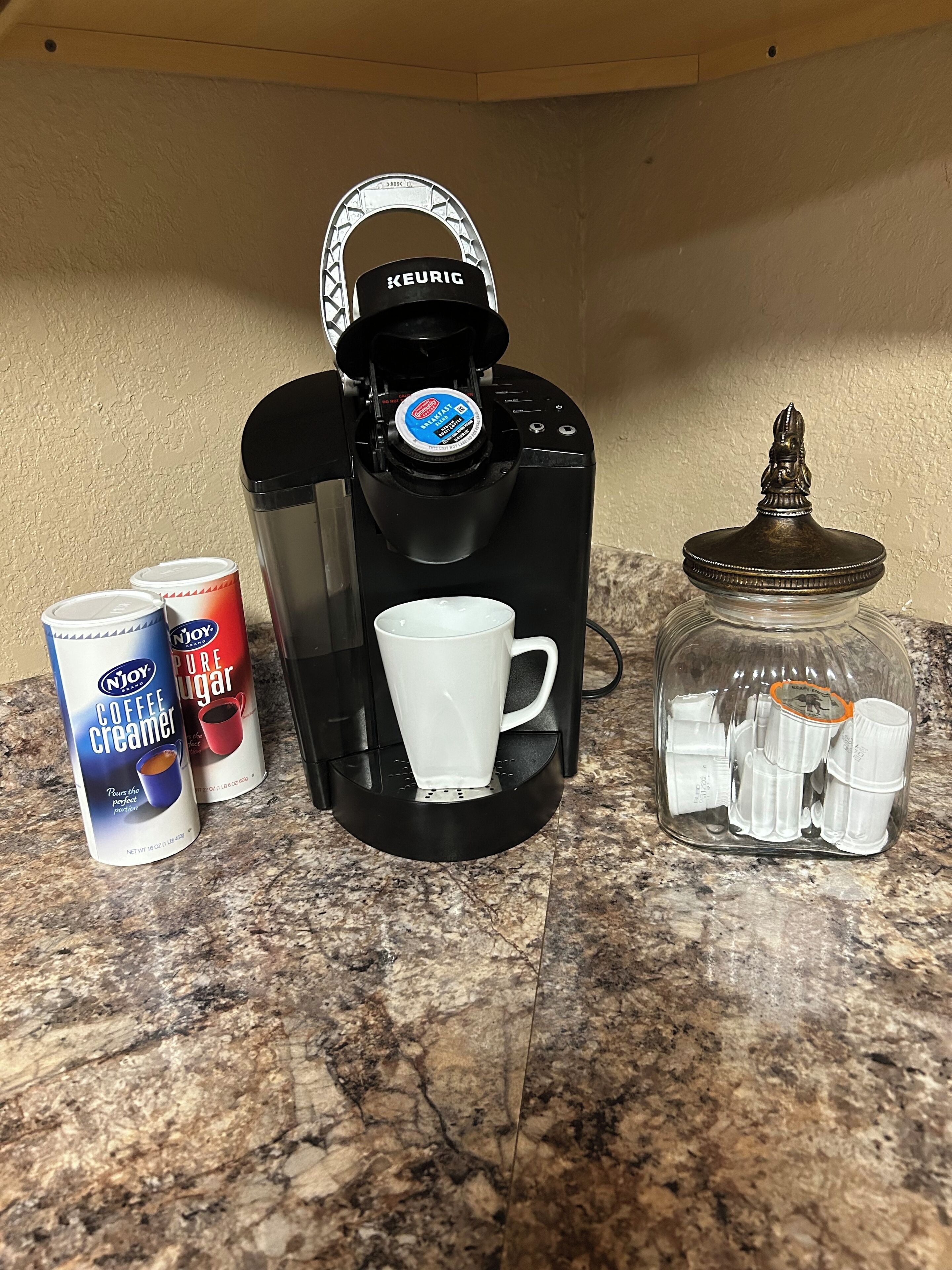 En Keurig kaffemaskine med en kaffekapsel, en hvid kop og to kaffemælkekander på en granitbordplade.