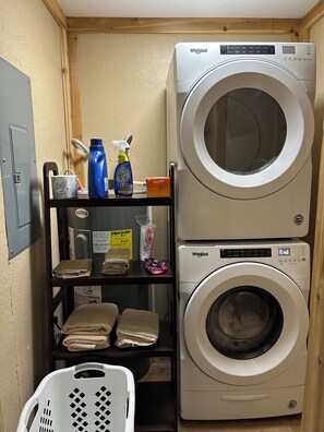 A laundry area with a stacked washer and dryer, a shelf with cleaning supplies, and a basket.