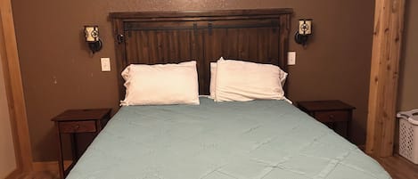 Une chambre comprenant un lit, une tête de lit en bois, un climatiseur fixé au mur, des tables de chevet et un panier à linge.