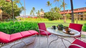 A patio with red chairs and a table, overlooking a lush garden and a building with a red roof. - Affordable quality in central location, Oceanside Complex  (Kihei)