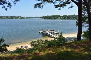 A lakeside view with a dock extending into the water, a sandy beach, and a grassy area with trees. - Beautiful waterfront home on Bass River (South Yarmouth)