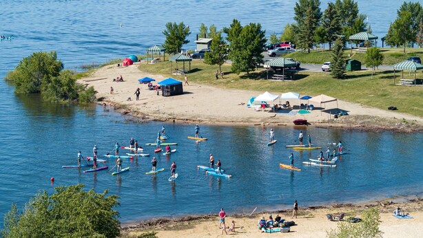 Ein Campingplatz am Seeufer mit Zelten, Picknicktischen und Stand-Up-Paddle-Boardern.