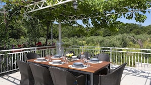 Ein Essbereich im Freien mit einem Tisch für sechs Personen, umgeben von Korbstühlen, unter einer Pergola mit Blick auf einen üppigen Garten und Bäume.