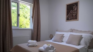 Ein ordentlich bezogenes Bett mit Handtüchern, ein Fenster mit Vorhängen, ein Bild an der Wand und ein Nachttisch mit einer Lampe.