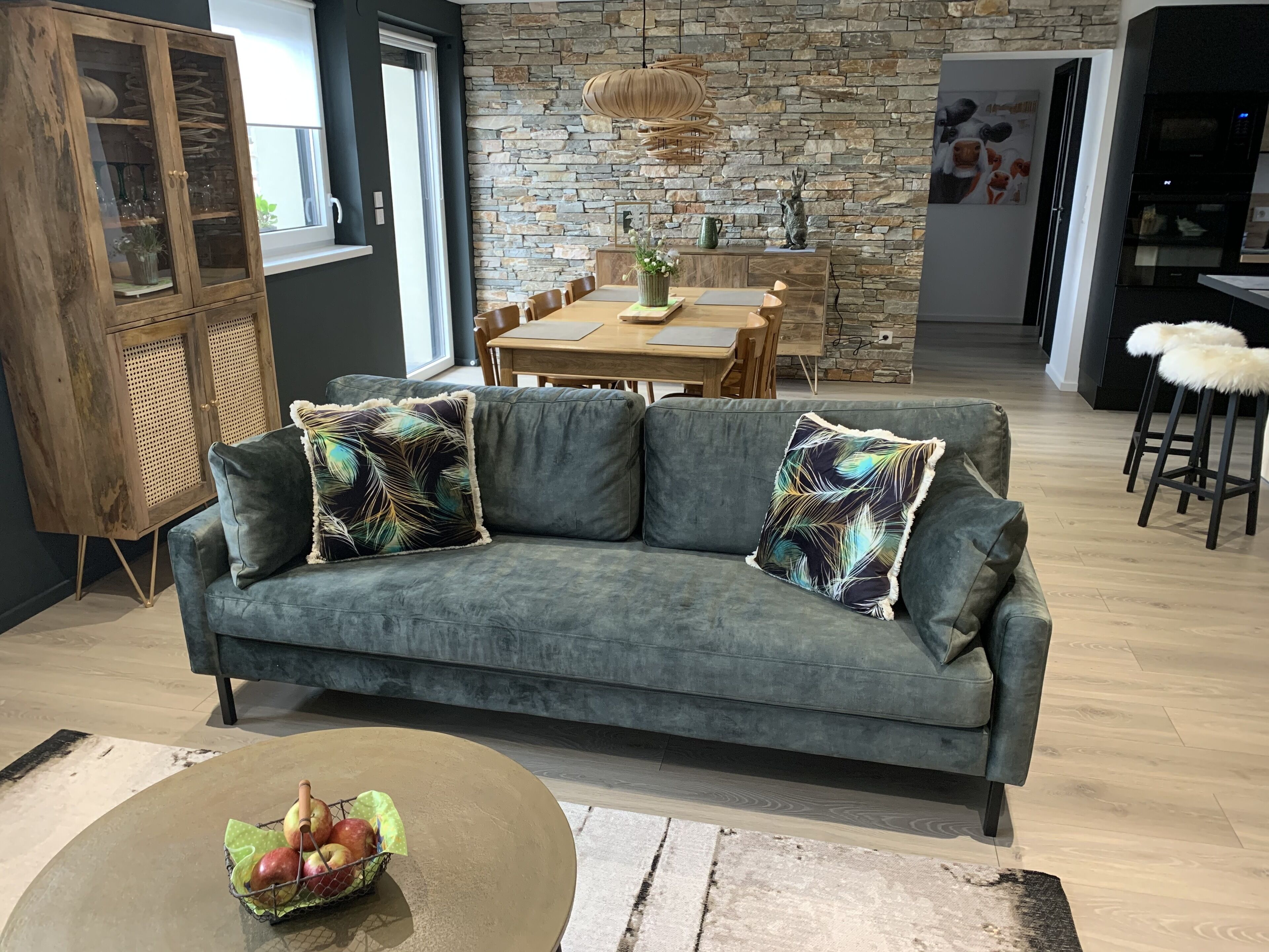 Un salon moderne avec un canapé gris, un meuble en bois et un coin repas agrémenté d’un mur en pierre et d’une table en bois.