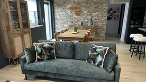 A modern living room with a grey sofa, a wooden cabinet, and a dining area with a stone wall and wooden table. - "LE SESAME" charming gite near Colmar (Wickerschwihr)