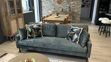 Un salon moderne avec un canapé gris, un meuble en bois et un coin repas agrémenté d’un mur en pierre et d’une table en bois.