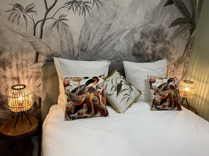 Una cama con ropa de cama blanca, dos almohadas con estampados de pájaros y dos lámparas fijadas a la pared.