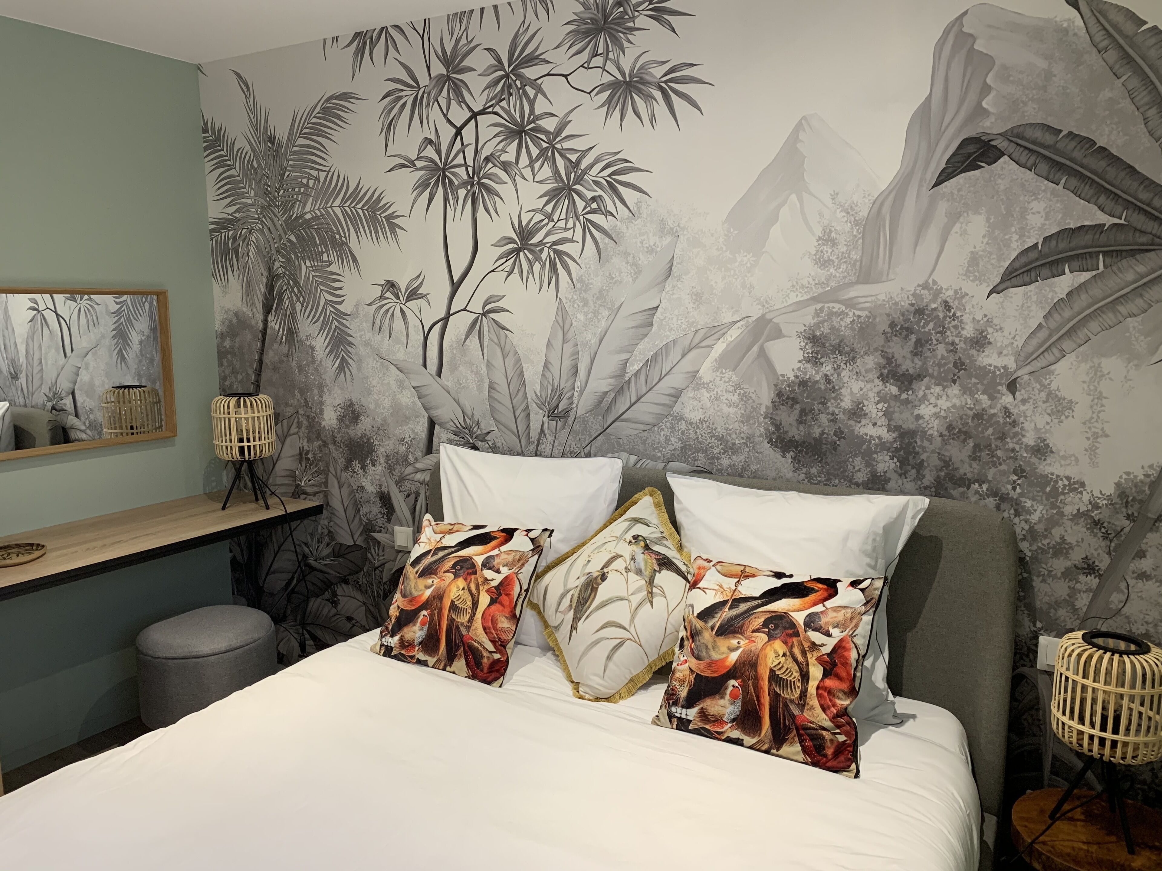 A bedroom with a large bed, two pillows with bird prints, a bedside table, a lamp, and a wall mural of bamboo and birds.