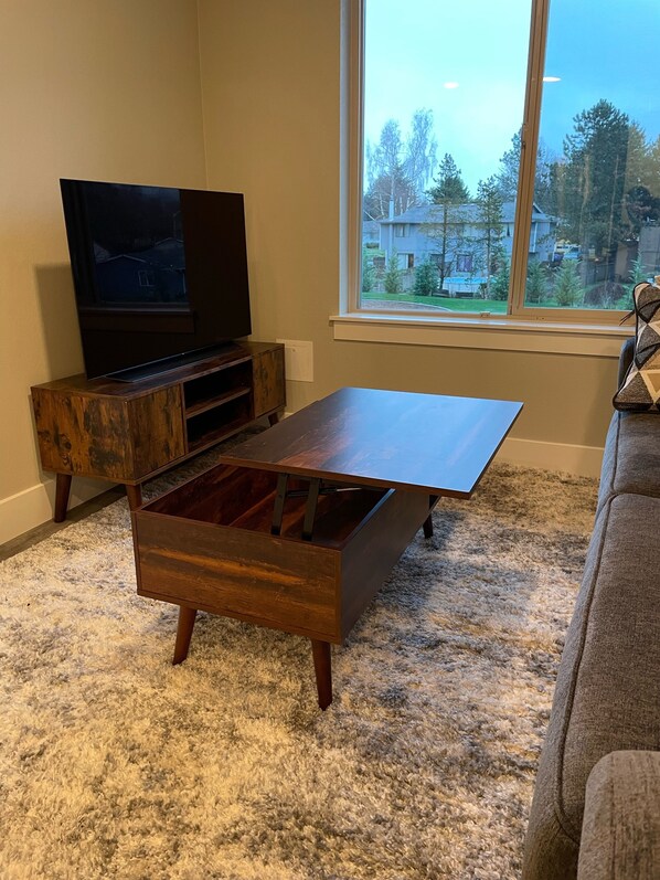 A living room with a wooden entertainment center, a flat-screen TV, a large window with a view of houses and trees, and a fluffy carpet.