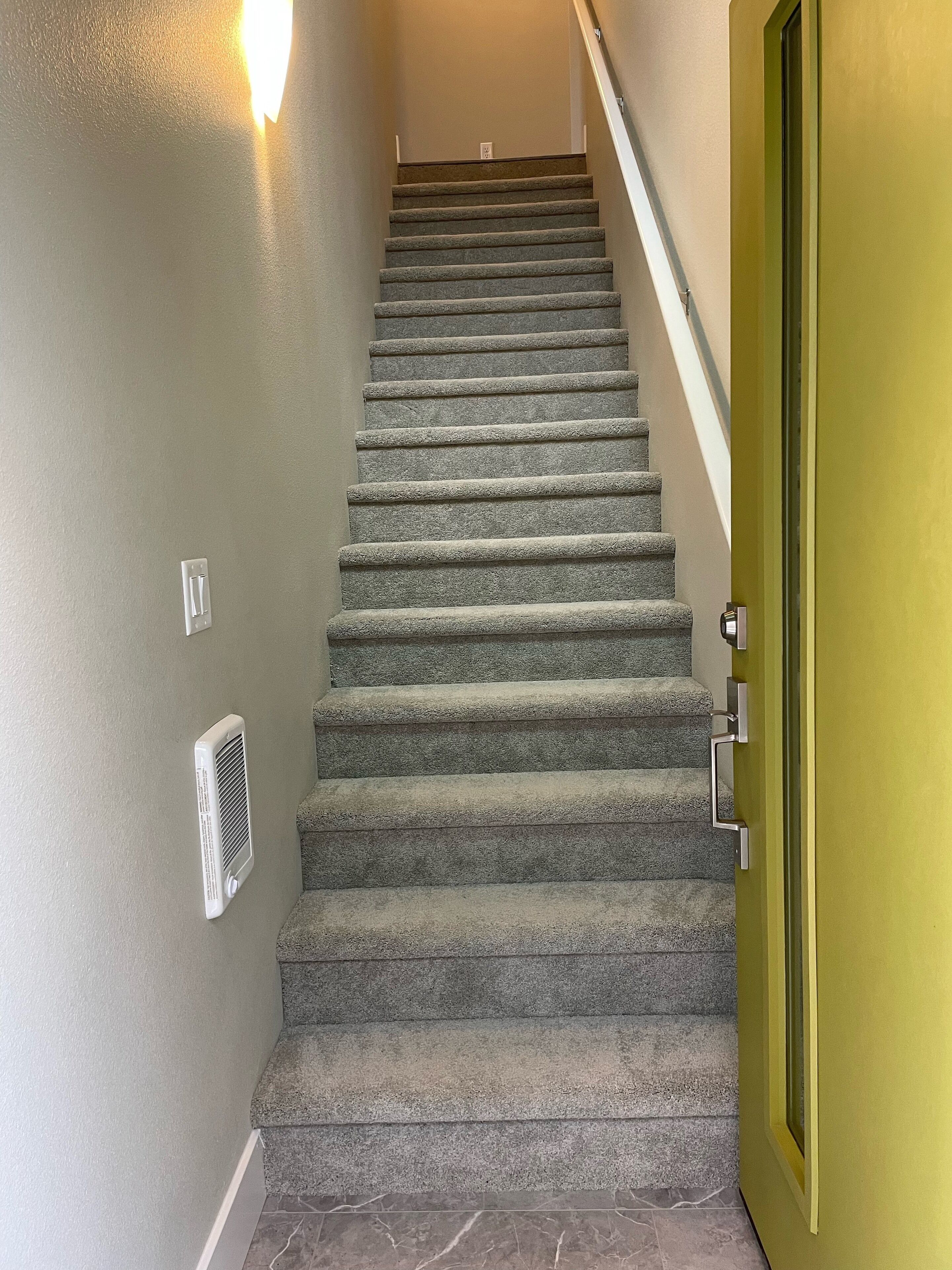 A carpeted staircase with a handrail, a wall-mounted vent, and a green door at the bottom.