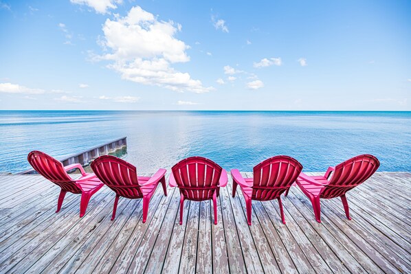 A wooden deck with five red chairs facing a calm sea under a clear sky. - Lexington Beach house, On the water with private beach (Lexington)