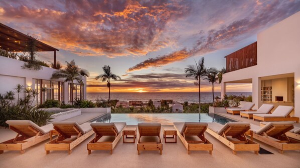 A poolside area with lounge chairs, a view of the ocean, and a sunset.
