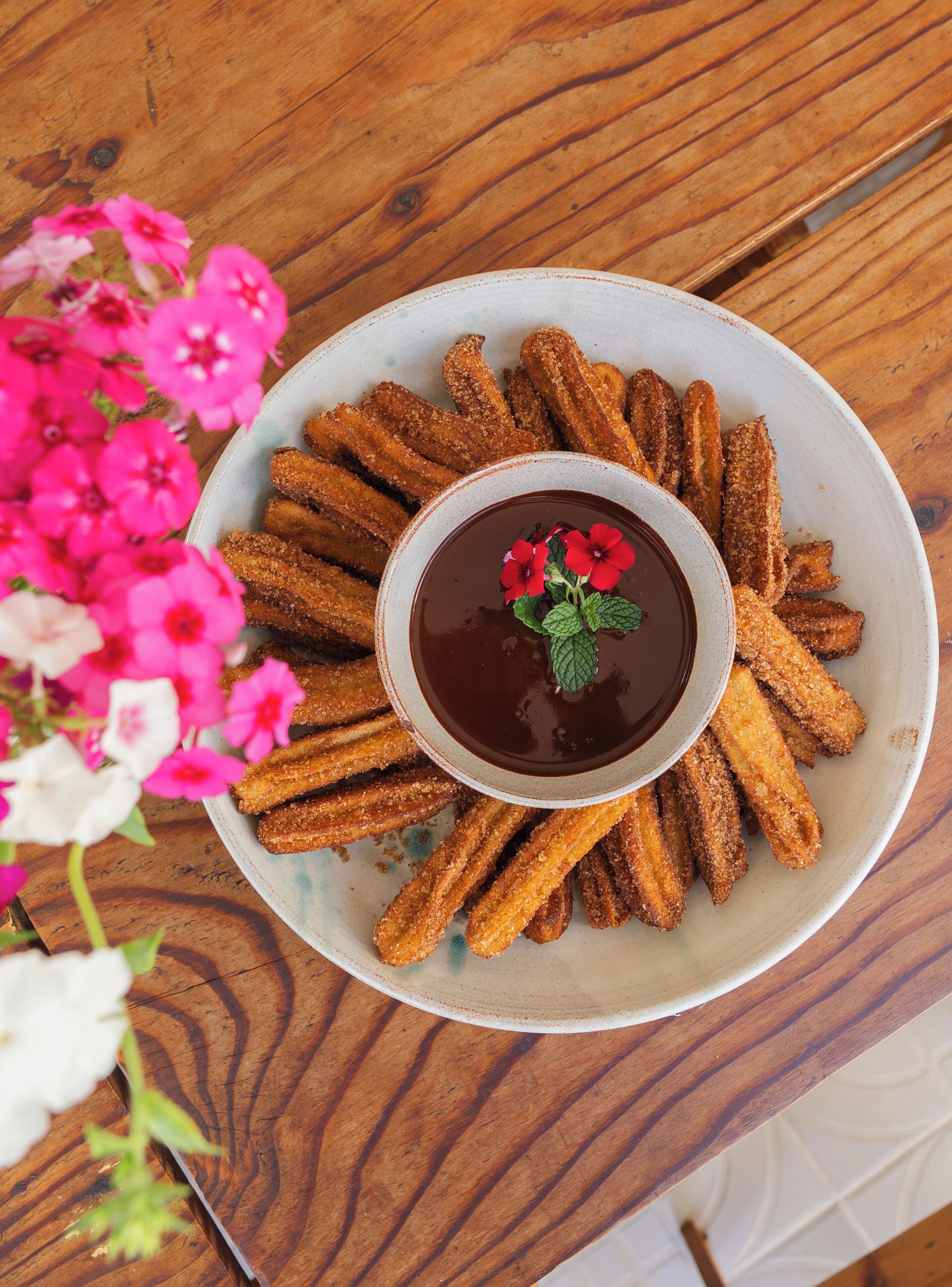 A plate of churros with a chocolate dipping sauce and a flower garnish, placed on a wooden table.