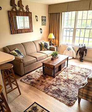 Un salon avec un canapé, une table basse et un tapis. Il y a une fenêtre avec des rideaux, une lampe et des éléments décoratifs sur les murs.