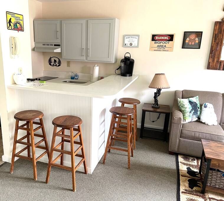 A compact living space with a kitchenette, a sofa, and a coffee table. There are three wooden stools near the kitchen counter.