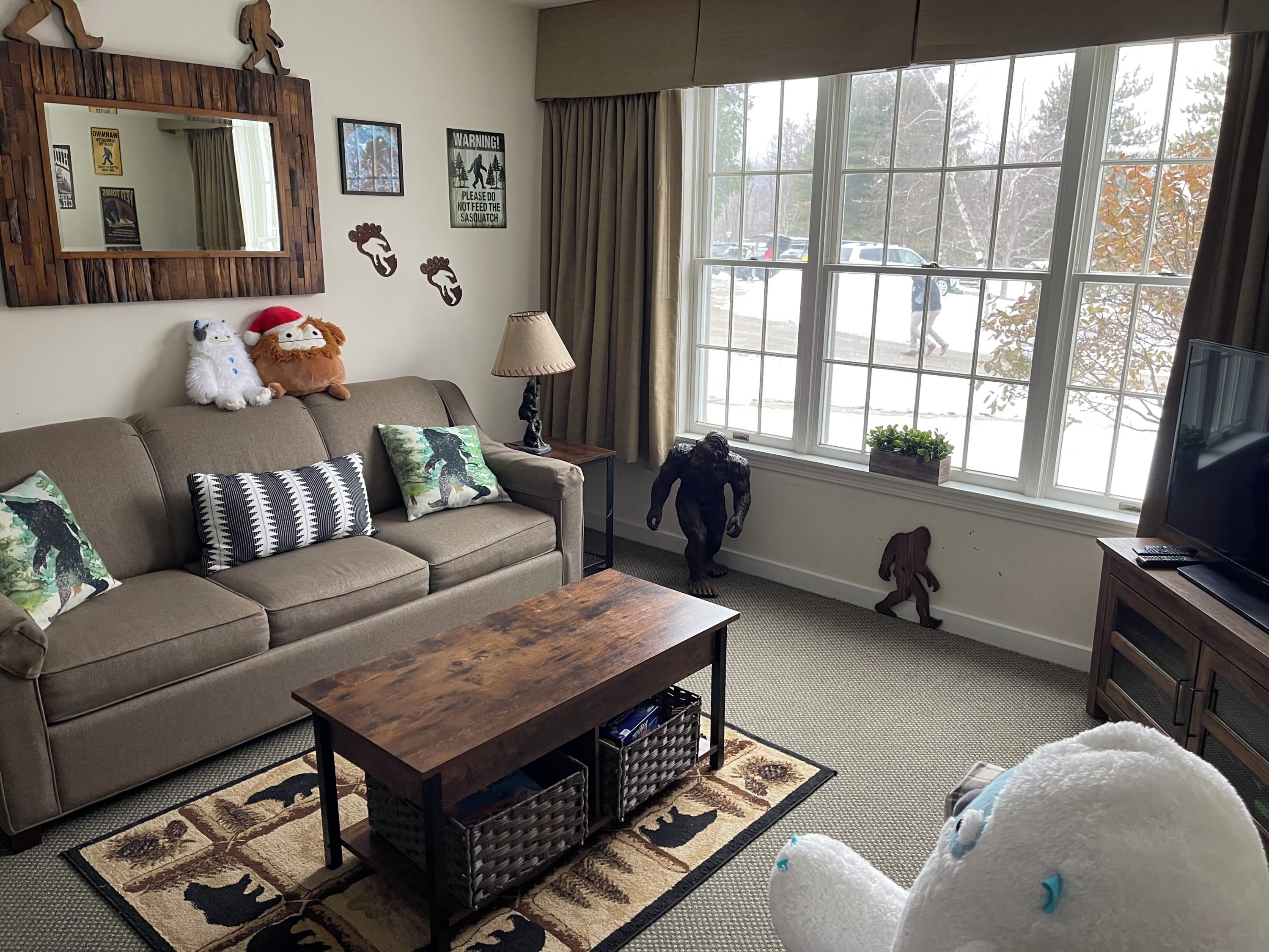 A living room with a sofa, a coffee table, a television, and a window with a view of parked cars.