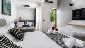 A modern bedroom with a large bed, a grey sofa, a TV, and a view of greenery outside. - Two Bedroom Villa with Private pool just 100m from the Beach (Heraklion)
