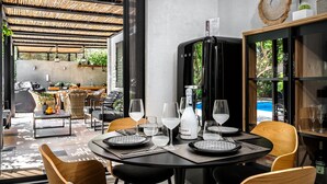 A dining area with a round table set for six, surrounded by wooden chairs. A black cabinet with glass doors is visible to the right, and a grill area is in the background. - Two Bedroom Villa with Private pool just 100m from the Beach (Heraklion)