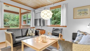 Ett mysigt vardagsrum med en soffa, fåtöljer, ett bord och en tv. Det finns fönster med gardiner, en hängande lampa och en tavla på väggen.