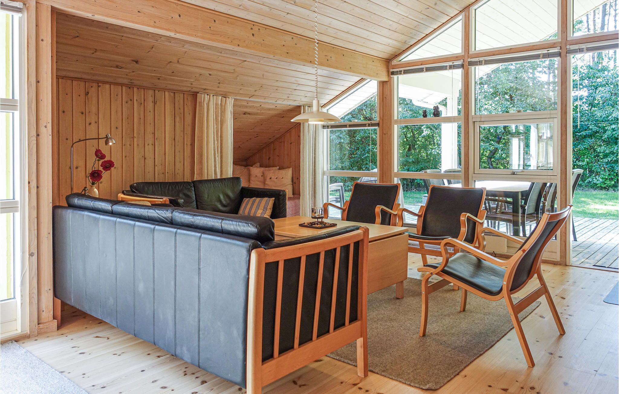 L’intérieur chaleureux d’un chalet avec un plafond en bois, un canapé, une table à manger avec des chaises et de grandes fenêtres donnant sur le paysage extérieur.