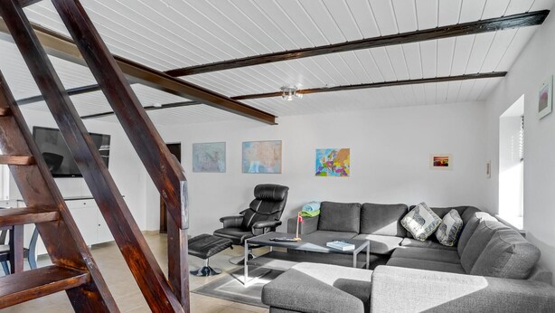 Un salon moderne avec un grand canapé d’angle gris, un fauteuil en cuir noir et une échelle en bois. Trois œuvres d’art encadrées ornent le mur, et un téléviseur y est fixé.