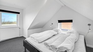 Une chambre moderne, aux murs blancs et au toit en pente, dotée d’une grande fenêtre, d’un lit recouvert de linge de maison blanc et d’une petite fenêtre donnant sur un mur de briques.
