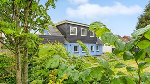 Une maison à deux étages, avec un toit sombre et des murs bleu clair, entourée d’une végétation luxuriante.