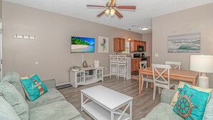 A living room with a sofa, coffee table, TV, and kitchen area with wooden cabinets and a dining table. - “Mermaid’s Dream” Short Walk from the Waves & Pool (Myrtle Beach)