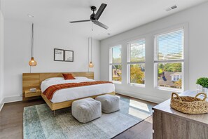 Une chambre à coucher moderne avec un lit en bois, deux poufs, un ventilateur de plafond et de grandes fenêtres.