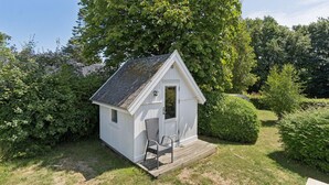 En liten vit träbod med ett sluttande tak, en dörr och ett fönster. Den står på en gräsyta, och på en träveranda framför boden finns en stol och ett litet bord.