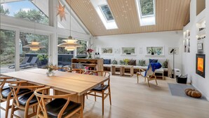 Un'ampia zona giorno con soffitto alto, travi in legno e grandi finestre che lasciano entrare la luce naturale. Si trova una zona pranzo con tavolo e sedie in legno, e un'area salotto con divano e cuscini.