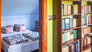 Un dormitori amb llit, almoinos i una tauleta de nit. A la dreta del llit hi ha una biblioteca plena de llibres.