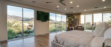 A spacious bedroom with a large bed, a ceiling fan, and a view of mountains through floor-to-ceiling windows.