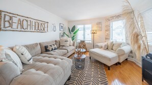 A living room with a beige sofa, a white armchair, and a coffee table with decorative items. A 'BEACHHOUSE' sign is on the wall, and there are windows with blinds.