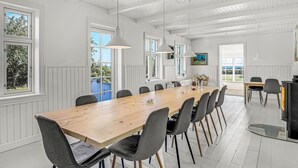 A spacious dining area with a long wooden table, grey upholstered chairs, pendant lighting, and large windows offering views of the outdoors. - 5 bedroom nice home in Svaneke (Svaneke)