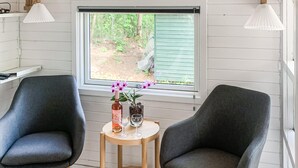 Et værelse med to mørkegrå lænestole, et lille træbord med en vase blomster, et vinglas og en flaske. Et vindue med udsigt til grøn bevoksning er synligt.