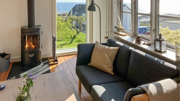 Un soggiorno accogliente con camino moderno, un divano confortevole, un tavolino con un vaso di fiori e vista mare attraverso ampie finestre.