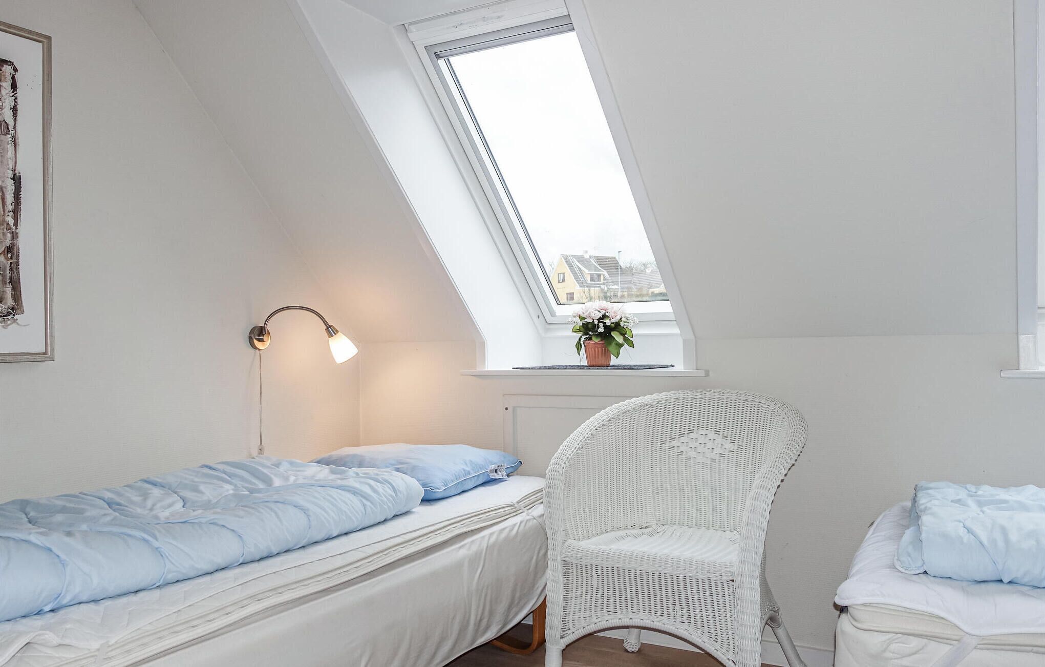 A small, white, single-room apartment with a sloped ceiling, a skylight, a white wicker chair, a bed with blue bedding, and a wall-mounted lamp.