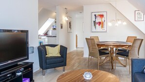 A modern living room with a flat-screen TV, a dark armchair, a round wooden coffee table, and a dining area with wicker chairs and a wooden table. - Stunning apartment in Gudhjem with WiFi (Gudhjem)